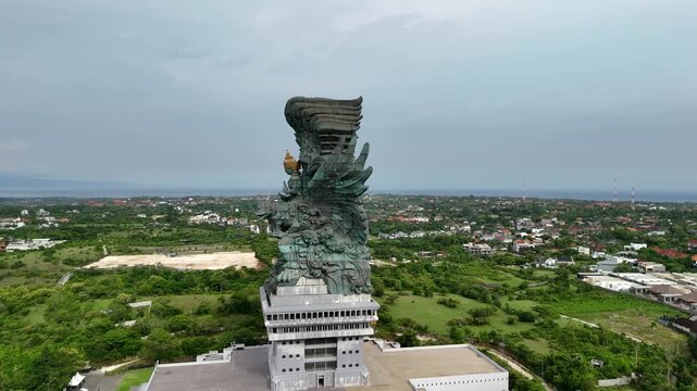 Drone approaching and passing very close to the huge bronze statue of Garuda Wisnu Kencana against Bali landscape background