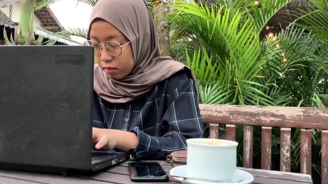 Selective focus on a wallet and saucer on a wooden table. In the blurred background, a Muslim woman in a hijab checks her smartphone and drinks coffee at an outdoor cafe. Relaxing lifestyle concept.