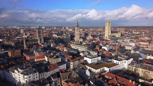 Cinematic aerial Ghent Belgium showcasing the stunning medieval cityscape with iconic belfry tower and Saint Bavo Cathedral rising above red-tiled rooftops under dramatic cloudy skies.