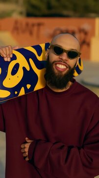 Smiling skateboarder portrait leaning on patterned yellow and blue deck in urban skatepark for vertical social banner and lifestyle background