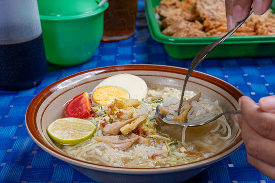 Hands using spoon and fork scooping up chicken soto soup served in a traditional indonesian warung setting