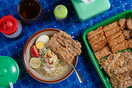 Overhead indonesian chicken soto soup with egg and tempeh tray served in traditional blue checkered warung setting