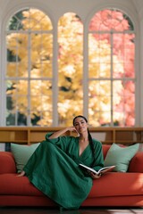 Naklejka premium Elegant woman reading on orange sofa in bright library with arched windows and autumn foliage for lifestyle background and editorial banner