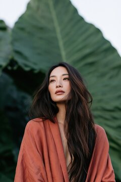 Vertical portrait of a woman draped in an orange robe against a giant tropical leaf background for editorial poster and wallpaper design