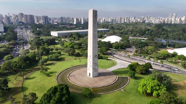 Aerial view of the towering Obelisk of Sao Paulo surrounded by lush green park trees against the distant city skyline.