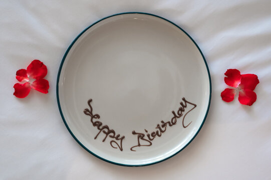 Happy birthday written in chocolate syrup on a white plate flanked by red rose petals.