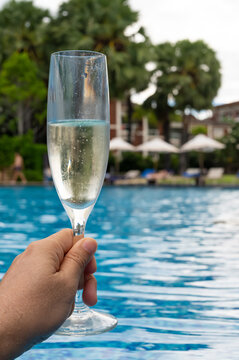 A hand holding a glass of champagne by a swimming pool.