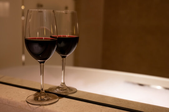 A glass of red wine with reflection by mirror at bathtub in a bathroom.