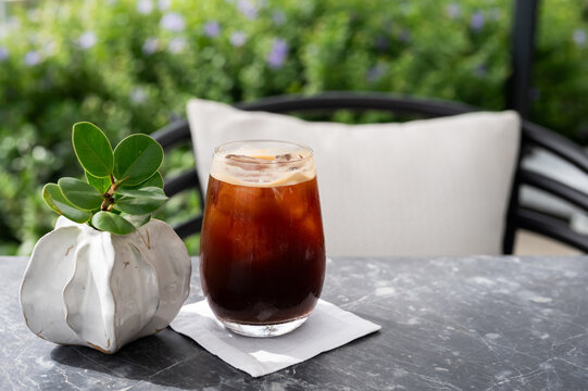 Glass of iced black coffee on a table with a small plant pot and garden view.