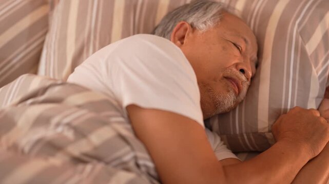 Elderly man shifts in his sleep and turns onto his side, settling deeper into the pillow in the quiet room. 4K retirement and healthcare b-roll with warm natural lighting.