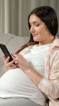 Expectant mother with a big belly resting on the sofa and using a smartphone for online shopping, preparing for childbirth and motherhood while enjoying a peaceful moment at home