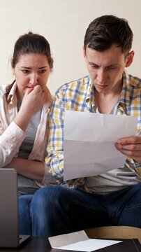 Worried young couple sitting on the couch and reading a letter with upsetting news. Stressed family holding a document about loan debt, eviction notice, or unpaid bills and feeling frustrated
