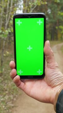 Man's hand holding a modern smartphone with a green screen and tracking markers while walking along a path in an autumn forest, a perfect mockup for navigation or app presentation