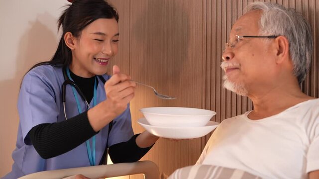 Cheerful nurse in blue scrubs raises a spoon toward an elderly man in glasses who leans forward to take a mouthful of soup. Ultra HD senior wellbeing footage from a contemporary care ward.