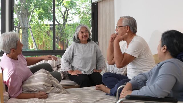 Four older adults hold an easy conversation across their beds with green garden foliage filling the window behind them. 4K senior healthcare footage shot in a modern Asian hospital room.