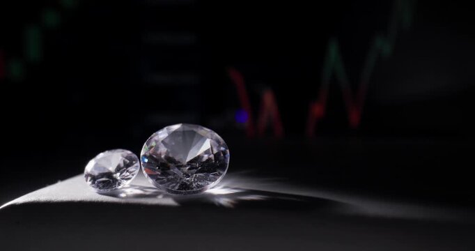 Closeup of two diamonds resting on dark desk with market chart on monitor, probe lens.