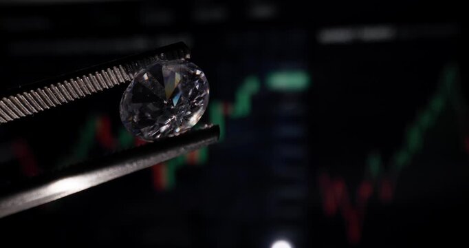 Diamond held by tweezers over trading monitor with market chart background, probe lens.