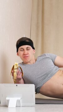 Funny lazy man in sportswear eating a banana while watching a workout video on a tablet instead of training, humorously depicting procrastination and the struggle with diet and fitness