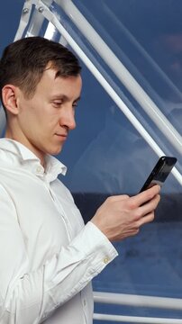 Young man in a white shirt concentrating on his smartphone while traveling, using an application to send a text message or browse online, with the sea visible through the window behind him