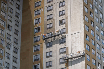 Workers fix the exterior of a tall building using lifts to apply materials to the surface in an urban setting during daylight hours © beholdereye