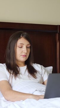 Beautiful brunette woman working on her laptop from the comfort of her bed, smiling while telecommuting, highlighting a modern, flexible, and comfortable work lifestyle