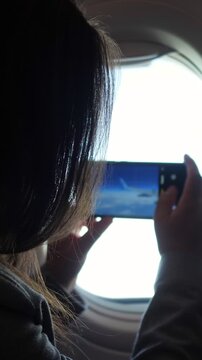 Young female traveler using her mobile phone to film the beautiful view of clouds and sky from the airplane window during a flight, creating content for social media and saving memories