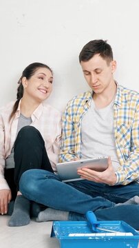 Young happy couple relaxing on the floor after painting a wall, planning their new apartment design on a tablet while discussing renovation ideas and choosing interior colors together