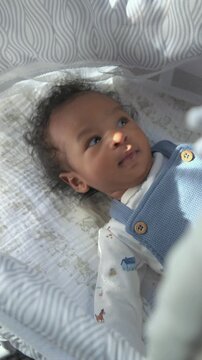 Vertical Screen: African American newborn baby smiles in a bassinet wearing a blue knit vest and animal print romper as a parent hand gently touches the chest in soft morning light.