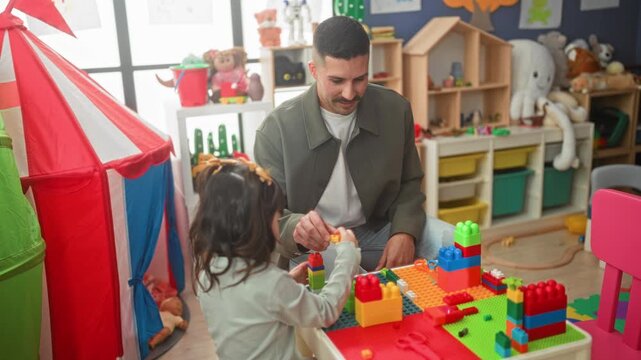 Man building colorful toy blocks with a little girl at a preschool playroom table, handing pieces and guiding construction; nurturing learning play.