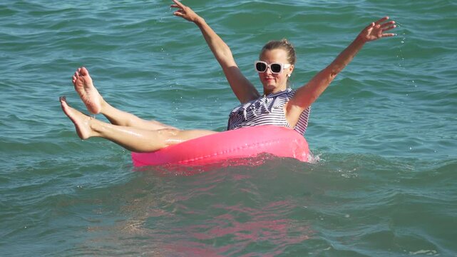 Beach lifestyle portrait featuring a cheerful woman floating on a pink tube in blue water. Steady medium shot captures joyful arm movements.