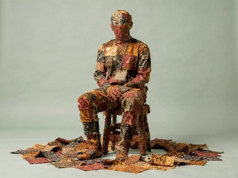 Seated human figure completely covered in textured batik textile scraps, a dense and handcrafted silhouette for slow fashion and craft content with copy space