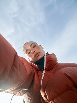Woman in a bright orange puffer jacket with a navy turtleneck, posing with a defiant expression against a cyan sky for fashion campaigns