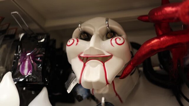 Halloween devil mask with red horns and eerie facial details hangs among costume accessories creating a spooky seasonal display inside a party store for shoppers at night