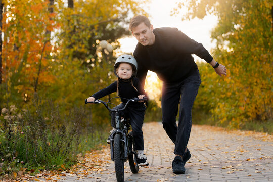 Father guides daughter in learning to ride bicycle on sunny autumn day, weekend time play with dad