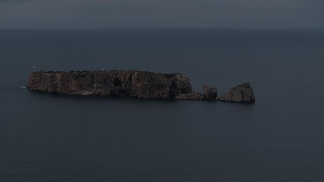 Aerial telephoto shot of the rocky Tsichli Baba islet with its sea cave and French monument, surrounded by the dark Ionian Sea at dusk in Pylos, Greece, creating a moody, isolated feeling.