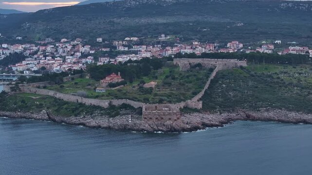 Aerial drone shot panning along the ancient stone walls of Niokastro fortress, revealing the Church of the Transfiguration, coastal harbour, and illuminated town of Pylos at twilight in Greece.