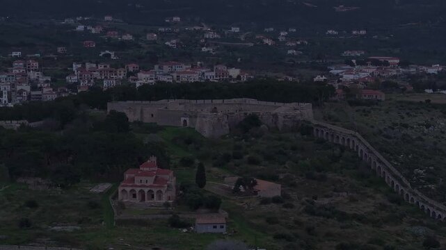 Aerial shot of Niokastro Castle, Church of the Transfiguration, and ancient aqueduct in Pylos, Greece, under a dark, moody twilight sky with town lights in the distance.