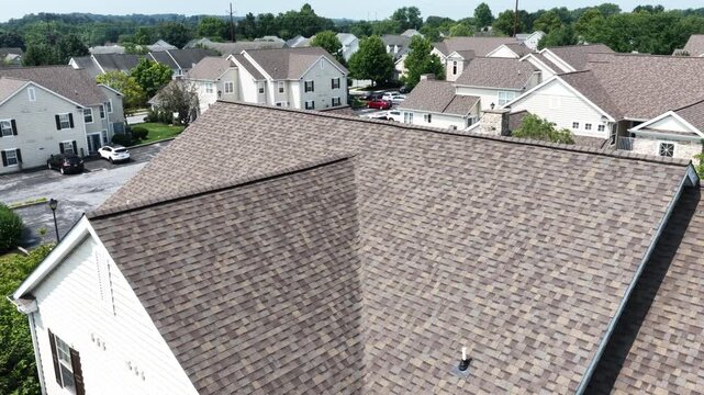 Steep asphalt shingle roof of townhome dominates frame beside development rooftops, driveways and mature trees spread across suburban USA neighborhood in Pennsylvania. Aerial drone orbit shot.