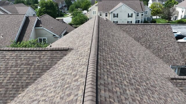 New asphalt shingle rooftop ridge overlooks townhome parking lot with white service van, houses and driveways fill quiet suburban American neighborhood below. Aerial drone flyover shot. Quiet scene.