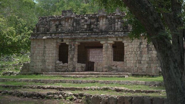 Static wide shot of an ancient Mayan stone building with columns framed by forest trees at Labna archaeological site in Yucatan, Mexico.