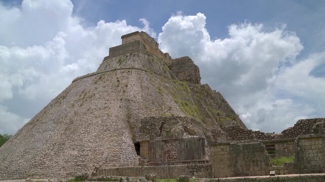 Static low angle wide shot of the iconic oval shaped Pyramid of the Magician at Uxmal Mayan ruins in Yucatan, Mexico under a cloudy blue sky.