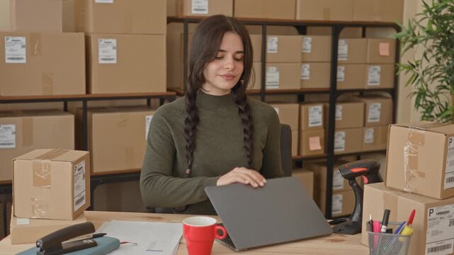 Young hispanic woman closing laptop beside stacked shipping boxes and barcode scanner at packing station in building; entrepreneurial confidence.