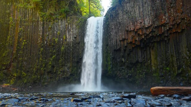 Waterfall beyond rocky foreground at Abiqua Falls, Oregon framed by towering basalt cliffs, low closeup as water rushes over smooth rocks