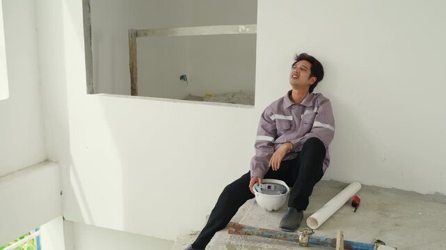 Construction inspector takes break sitting concrete floor inside unfinished building holding safety helmet resting against white wall calm moment with worker indoor building site safety equipment