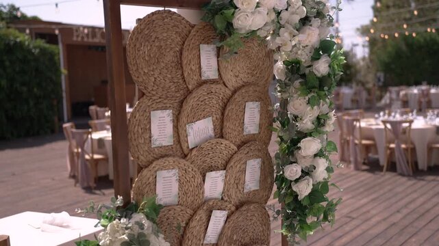 Decorations are arranged in an outdoor space for a wedding. There are tables set up and a display of names on woven circles with flowers around them.
