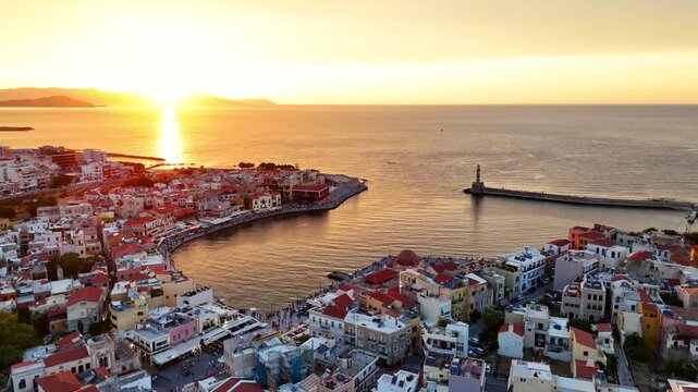 Chania Old Venetian Harbor and Egyptian Lighthouse glow at sunset in Chania, Crete, Greece, with dense historic waterfront buildings surrounding the harbor during a drone forward flyover.