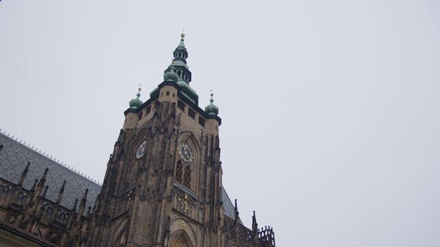 Great south tower of a gothic cathedral against a pale sky, clock faces, green copper cupola and gilded detail above a patterned slate roof and stone pinnacles.