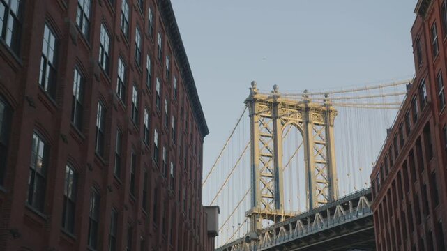 Lit steel bridge tower and suspension cables fill the gap between two red brick buildings, warm evening sun catching the ironwork above a corner sign.