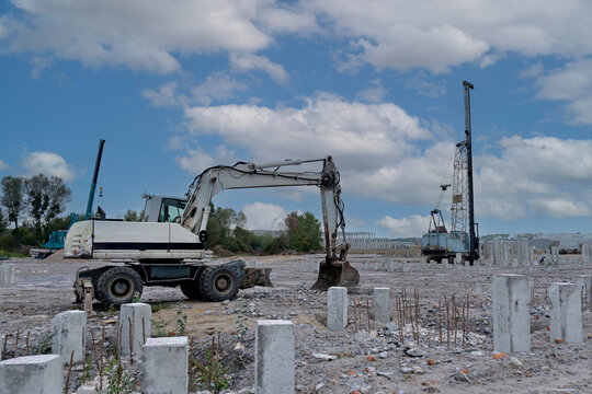 Wide view of a construction field with a white wheeled excavator in the foreground and a tall pile driver rig working on concrete foundation piles.