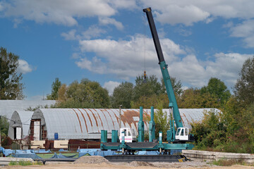 Heavy green mobile pile driver machine standing on a gravel base at a construction site with industrial hangars and green trees under sky. © Констянтин Батыльчук